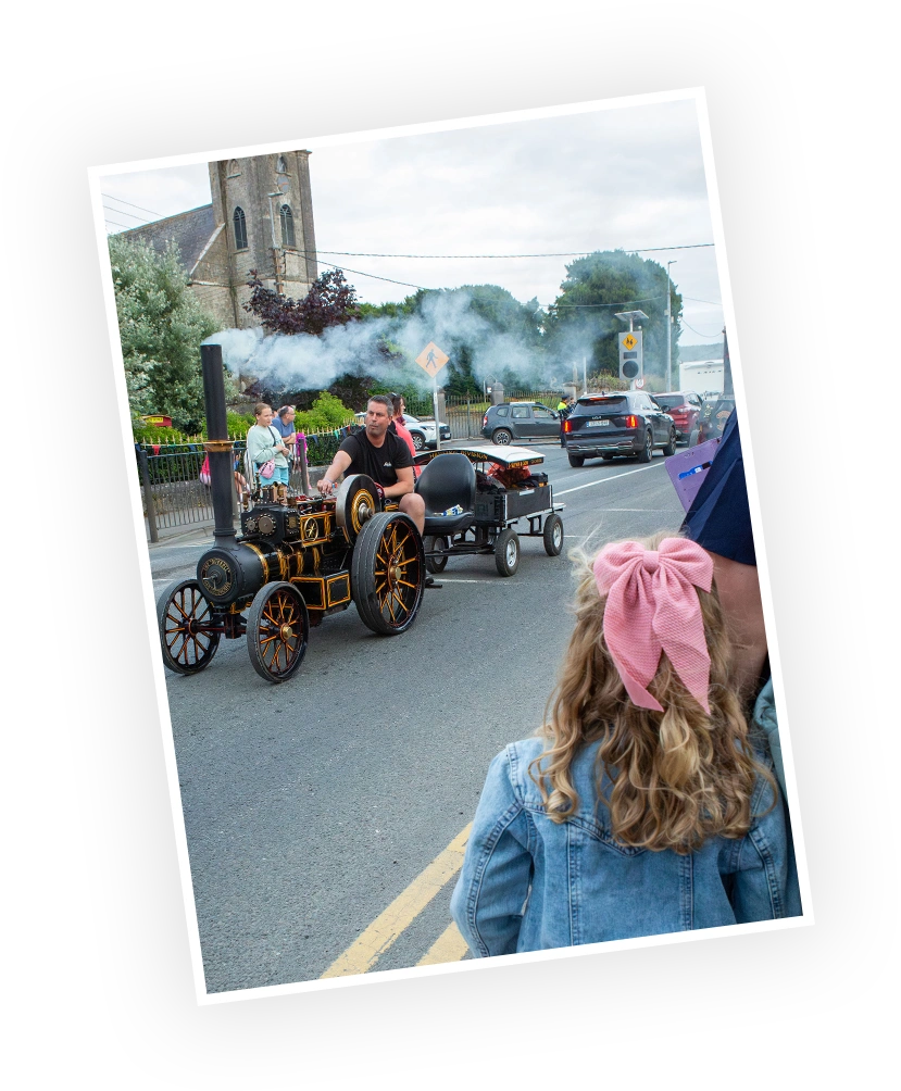 A vintage steam engine parades along a street during a local event, with a man operating the vehicle and children watching in excitement.