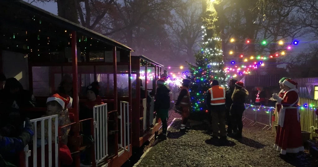Families enjoying a festive event outdoors, with a decorated Christmas tree and colourful lights illuminating the scene. Children wear Santa hats while boarding a festive train, and staff in holiday attire interact with visitors amidst a charming winter atmosphere.
