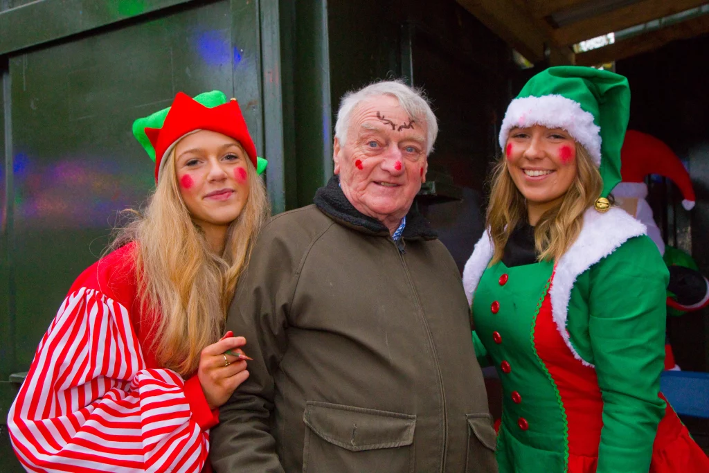 Two women dressed as festive elves pose with an older man in a green coat. Theyβre smiling and have playful red face paint, surrounded by Christmas decorations.