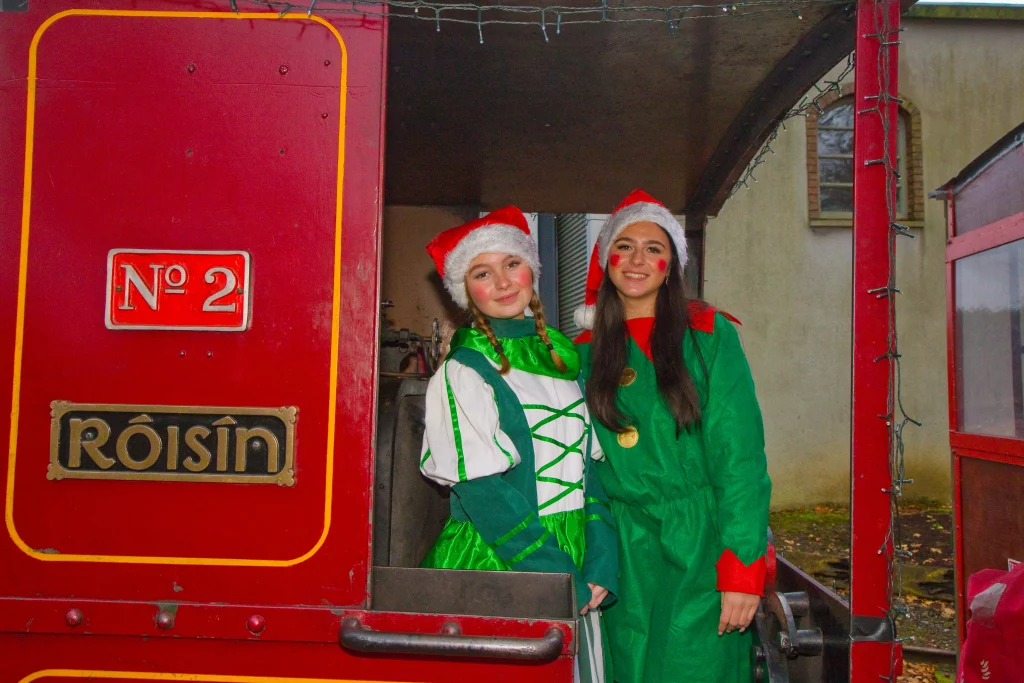 Two women in festive elf costumes pose beside a vintage train carriage, adorned with Christmas decorations.