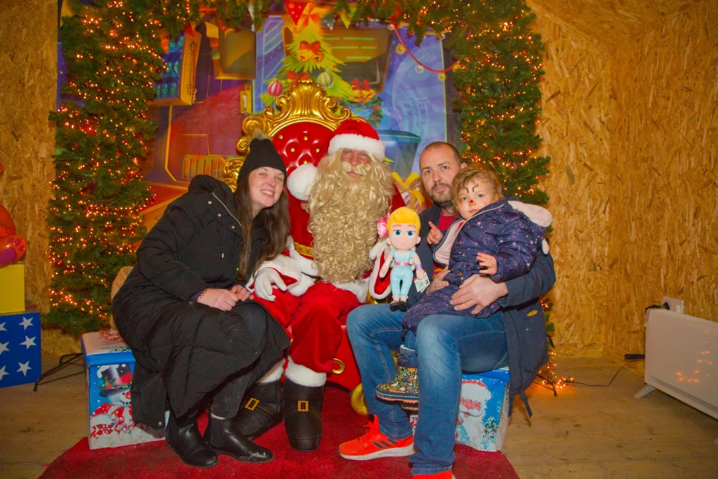 Family posing with Santa Claus in a festive setting, surrounded by Christmas decorations and twinkling lights, capturing a joyful holiday moment.